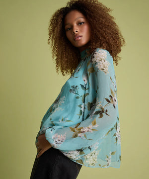 A woman with curly brown hair models a light blue floral long-sleeve blouse with black jeans against a solid light olive green background.