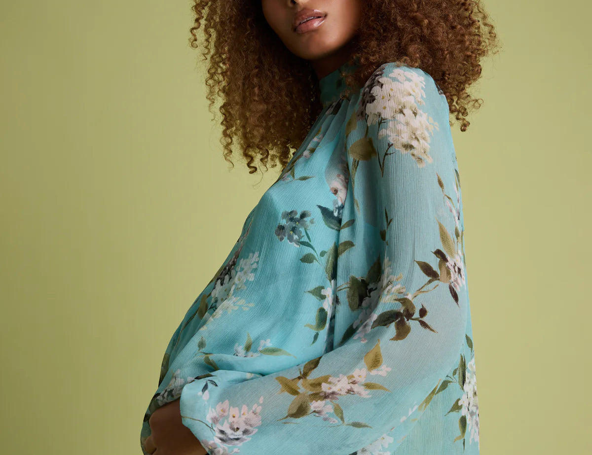A woman with curly brown hair models a light blue floral long-sleeve blouse with black jeans against a solid light olive green background.
