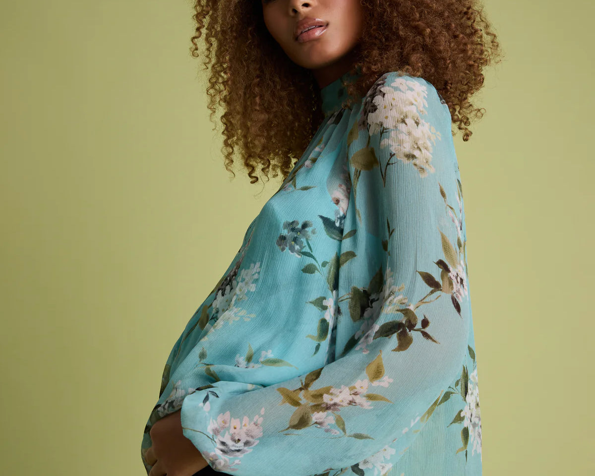 A woman with curly brown hair models a light blue floral long-sleeve blouse with black jeans against a solid light olive green background.