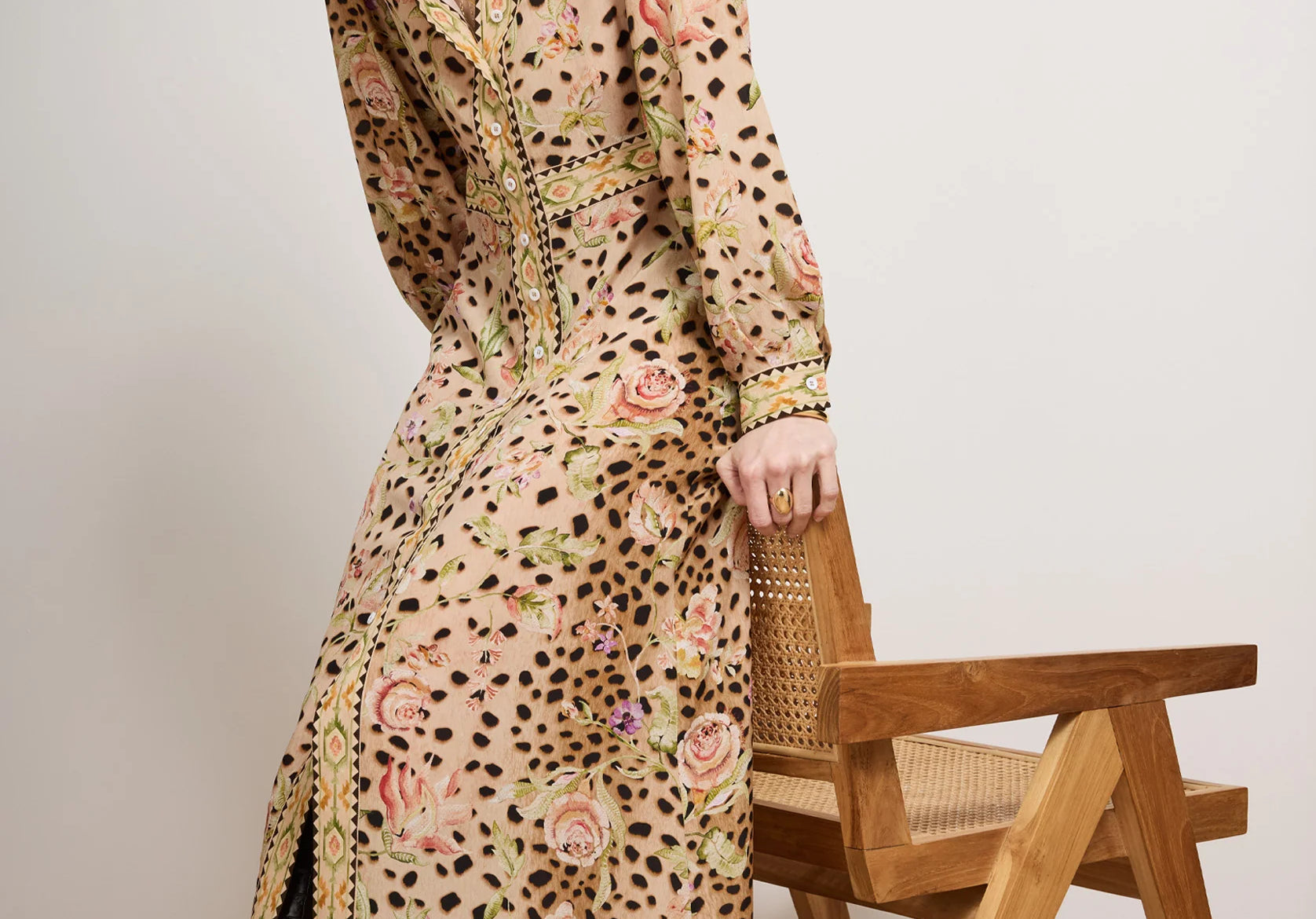 Woman wearing a long, patterned dress standing next to a wooden chair against a plain background