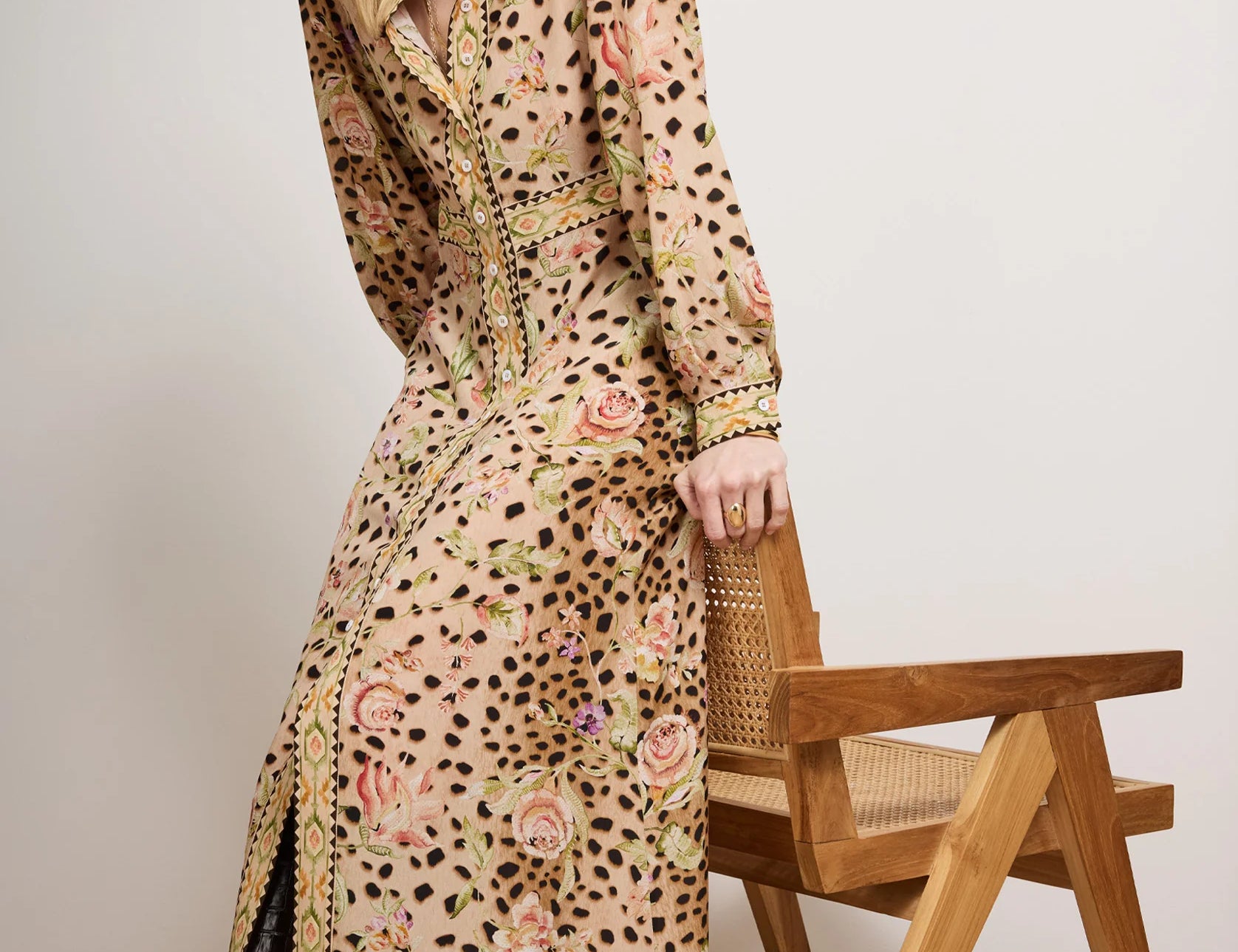 Woman wearing a long, patterned dress standing next to a wooden chair against a plain background