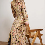 Woman wearing a long, patterned dress standing next to a wooden chair against a plain background
