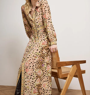 Woman wearing a long, patterned dress standing next to a wooden chair against a plain background