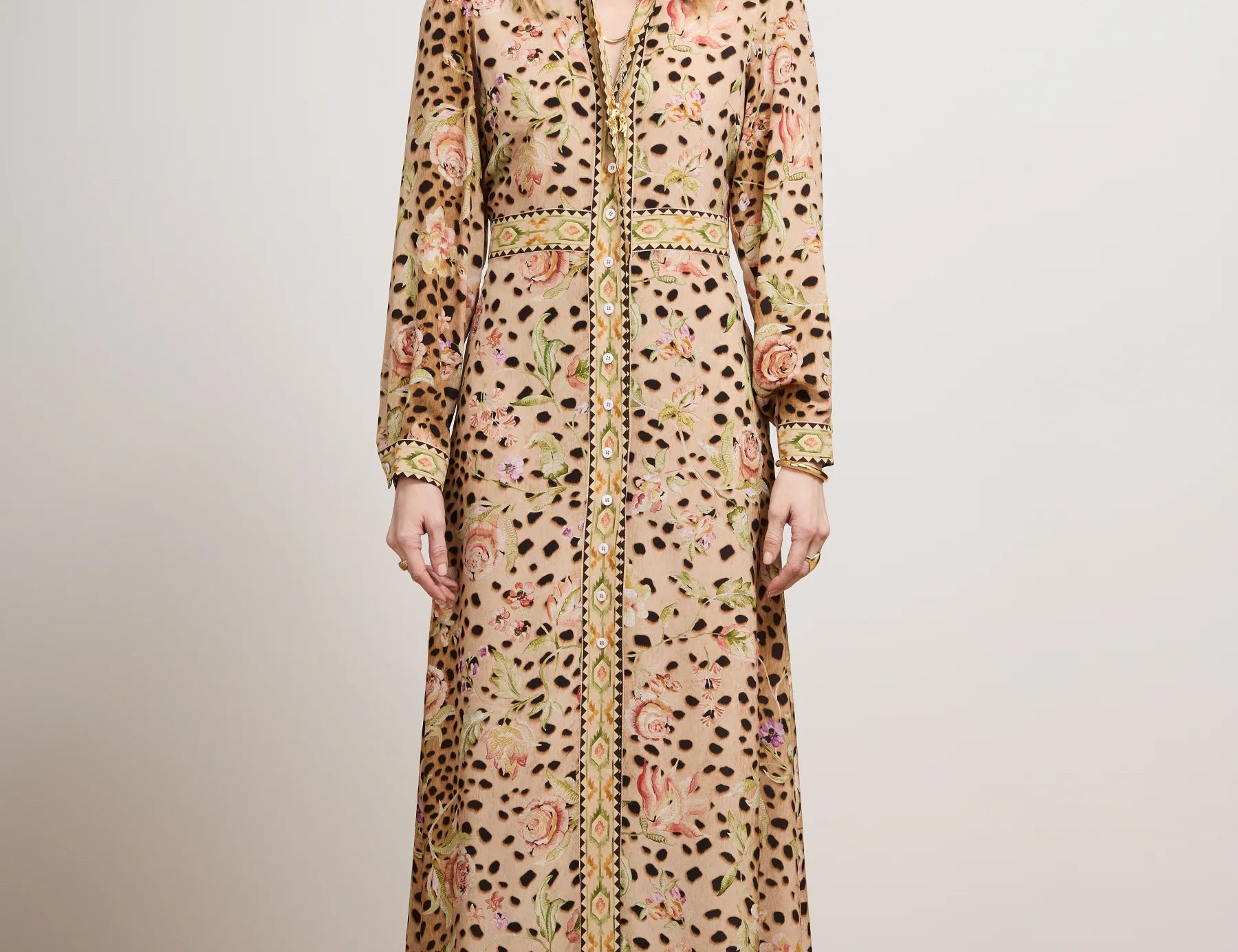 A model stands in a studio wearing a long-sleeved maxi shirt dress with a beige base, black leopard spots, and a pink and green floral pattern, paired with a matching belt and black shoes.