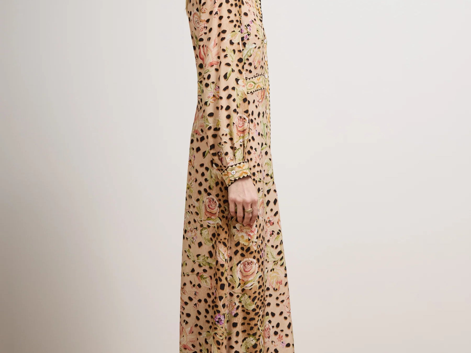 A woman stands in profile wearing a long-sleeved maxi dress with an all-over leopard print and floral embroidery in muted pink, green, and gold tones. She is photographed in a minimalist studio setting with a light beige wall and a light wood parquet floor.