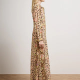 A woman stands in profile wearing a long-sleeved maxi dress with an all-over leopard print and floral embroidery in muted pink, green, and gold tones. She is photographed in a minimalist studio setting with a light beige wall and a light wood parquet floor.