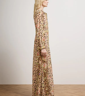 A woman stands in profile wearing a long-sleeved maxi dress with an all-over leopard print and floral embroidery in muted pink, green, and gold tones. She is photographed in a minimalist studio setting with a light beige wall and a light wood parquet floor.