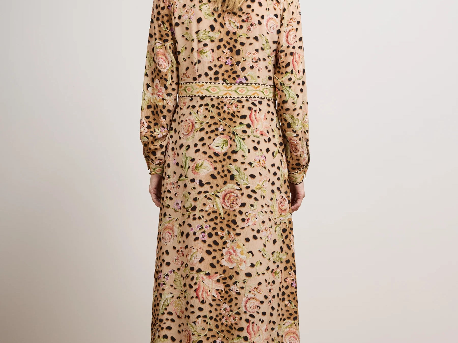 A woman viewed from behind wearing a long-sleeved maxi dress with an abstract floral and spotted pattern in beige, pink, green, and black, standing in a studio with a light gray wall and wood floor.
