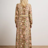 A woman viewed from behind wearing a long-sleeved maxi dress with an abstract floral and spotted pattern in beige, pink, green, and black, standing in a studio with a light gray wall and wood floor.