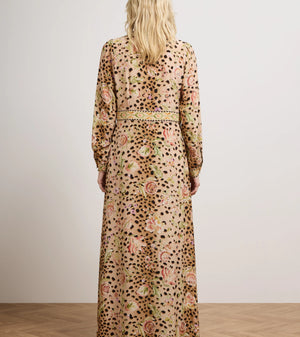 A woman viewed from behind wearing a long-sleeved maxi dress with an abstract floral and spotted pattern in beige, pink, green, and black, standing in a studio with a light gray wall and wood floor.