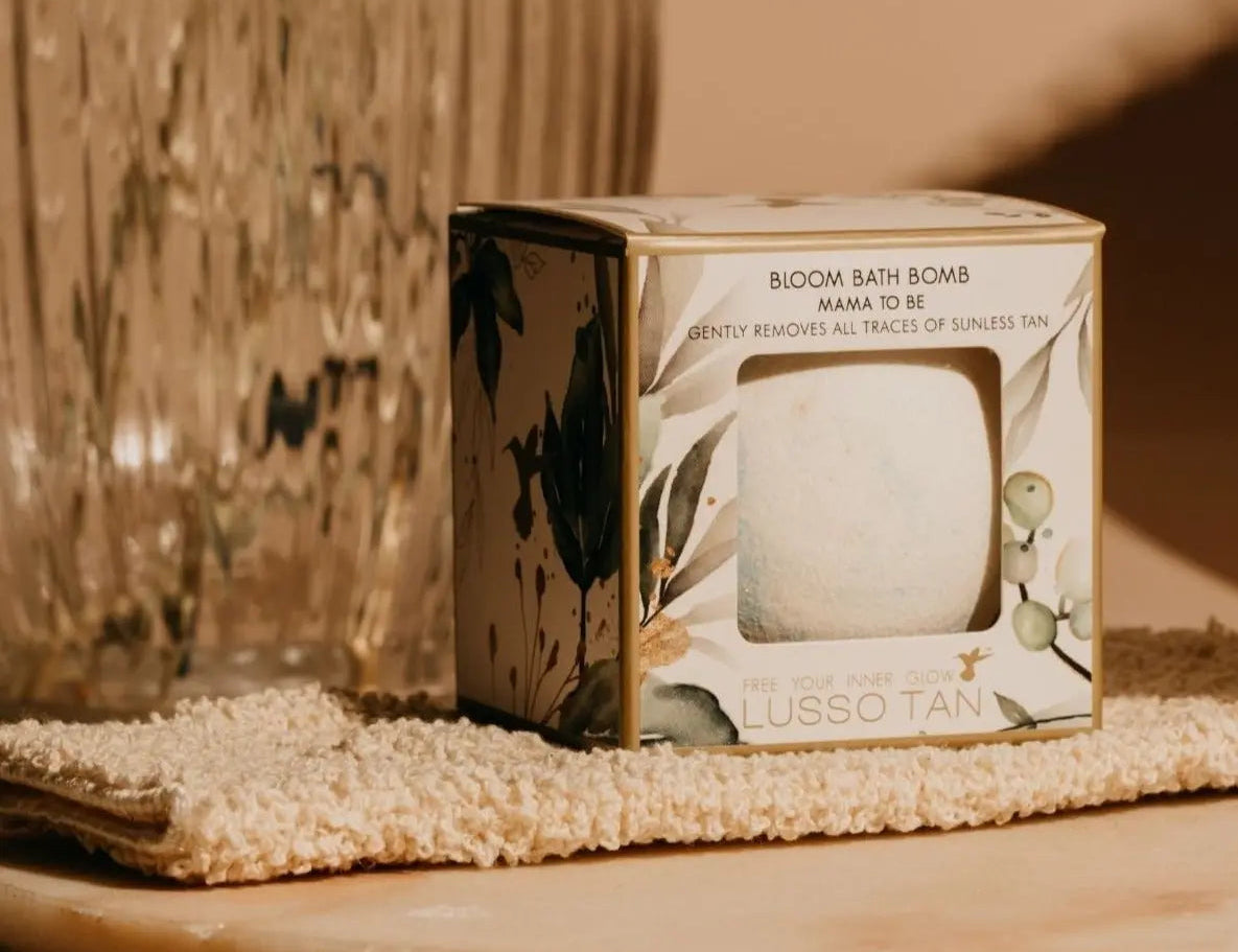 Tan Removing Bath Bomb Bloom in its boxed packaging, standing on a woven mitt beside a ribbed glass vase
