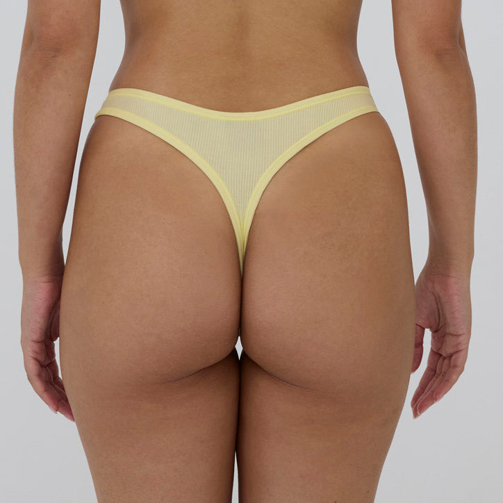 Rear view of a model wearing a pale yellow mesh thong with solid yellow trim, shown against a light gray studio background.