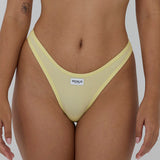 A close-up of a model wearing a pale yellow ribbed mesh thong with a white brand label reading 'RENUE' on the front, set against a solid gray background.
