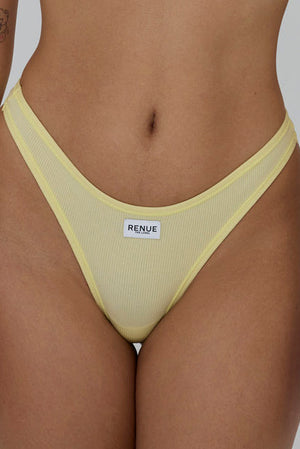 A close-up of a model wearing a pale yellow ribbed mesh thong with a white brand label reading 'RENUE' on the front, set against a solid gray background.
