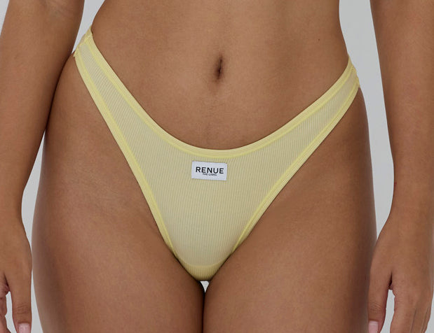 A close-up of a model wearing a pale yellow ribbed mesh thong with a white brand label reading 'RENUE' on the front, set against a solid gray background.