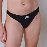 A close-up of a model wearing a black ribbed bikini bottom with a small white brand label reading 'RENUE' on the front, against a plain light gray background.