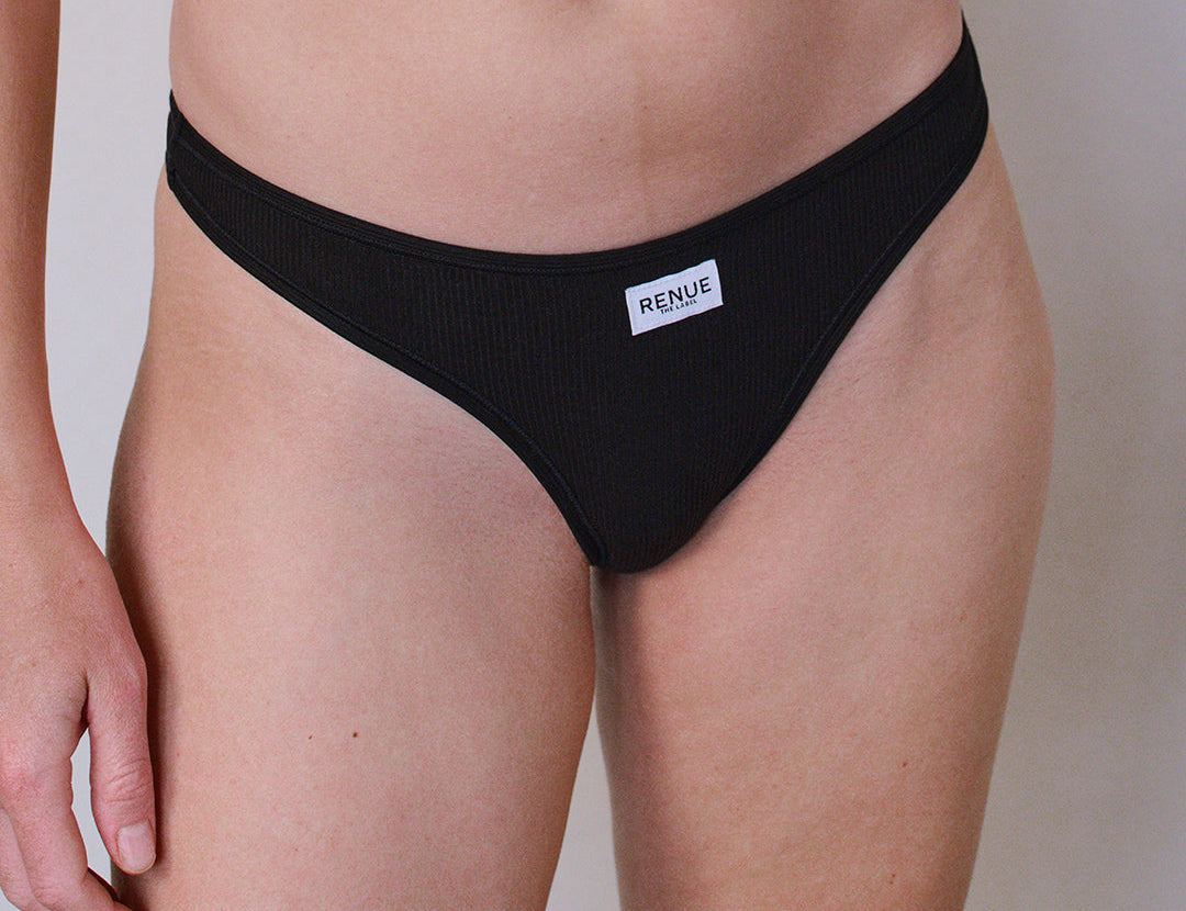 A close-up of a model wearing a black ribbed bikini bottom with a small white brand label reading 'RENUE' on the front, against a plain light gray background.