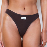 A close-up of a model wearing a dark brown ribbed bikini bottom with a white rectangular brand label that reads 'RENUE'.