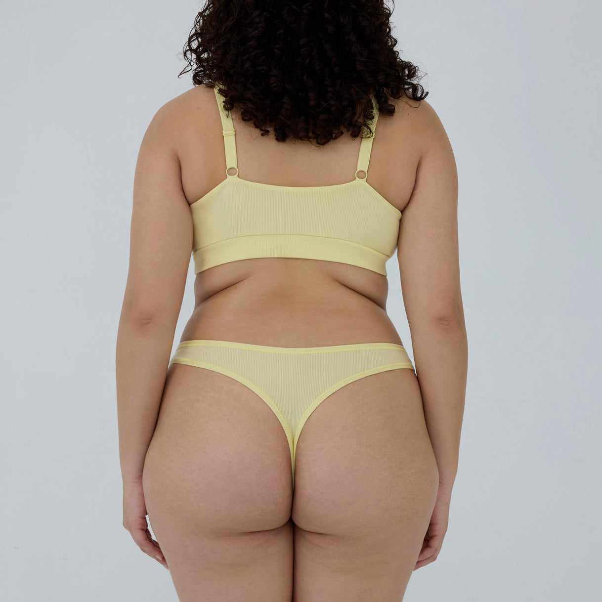 A model shown from behind wearing a pale yellow two-piece lingerie set, consisting of a bandeau-style bra and matching thong, against a plain light gray background.