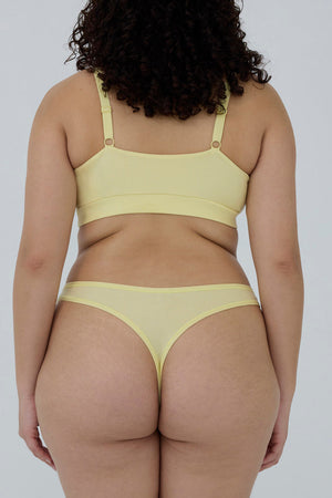 A model shown from behind wearing a pale yellow two-piece lingerie set, consisting of a bandeau-style bra and matching thong, against a plain light gray background.