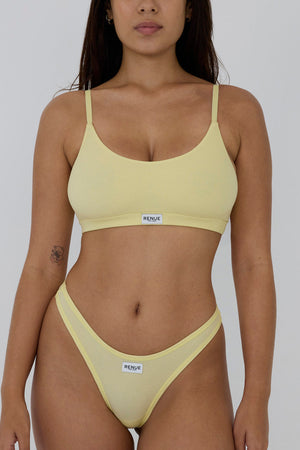 A woman modeling a pale yellow two-piece bikini set with a scoop-neck bra top and matching high-cut bottoms, featuring a small white brand label that reads 'MELUSIN' on the front of both pieces, against a plain light gray studio background.