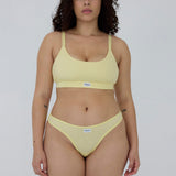 A woman modeling a pale yellow two-piece bikini set against a plain light gray studio background. The set includes a scoop-neck bikini top with thin shoulder straps and matching low-rise bikini bottoms.