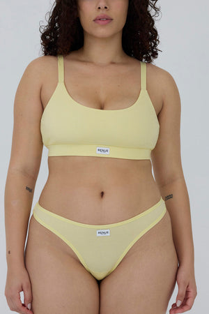 A woman modeling a pale yellow two-piece bikini set against a plain light gray studio background. The set includes a scoop-neck bikini top with thin shoulder straps and matching low-rise bikini bottoms.