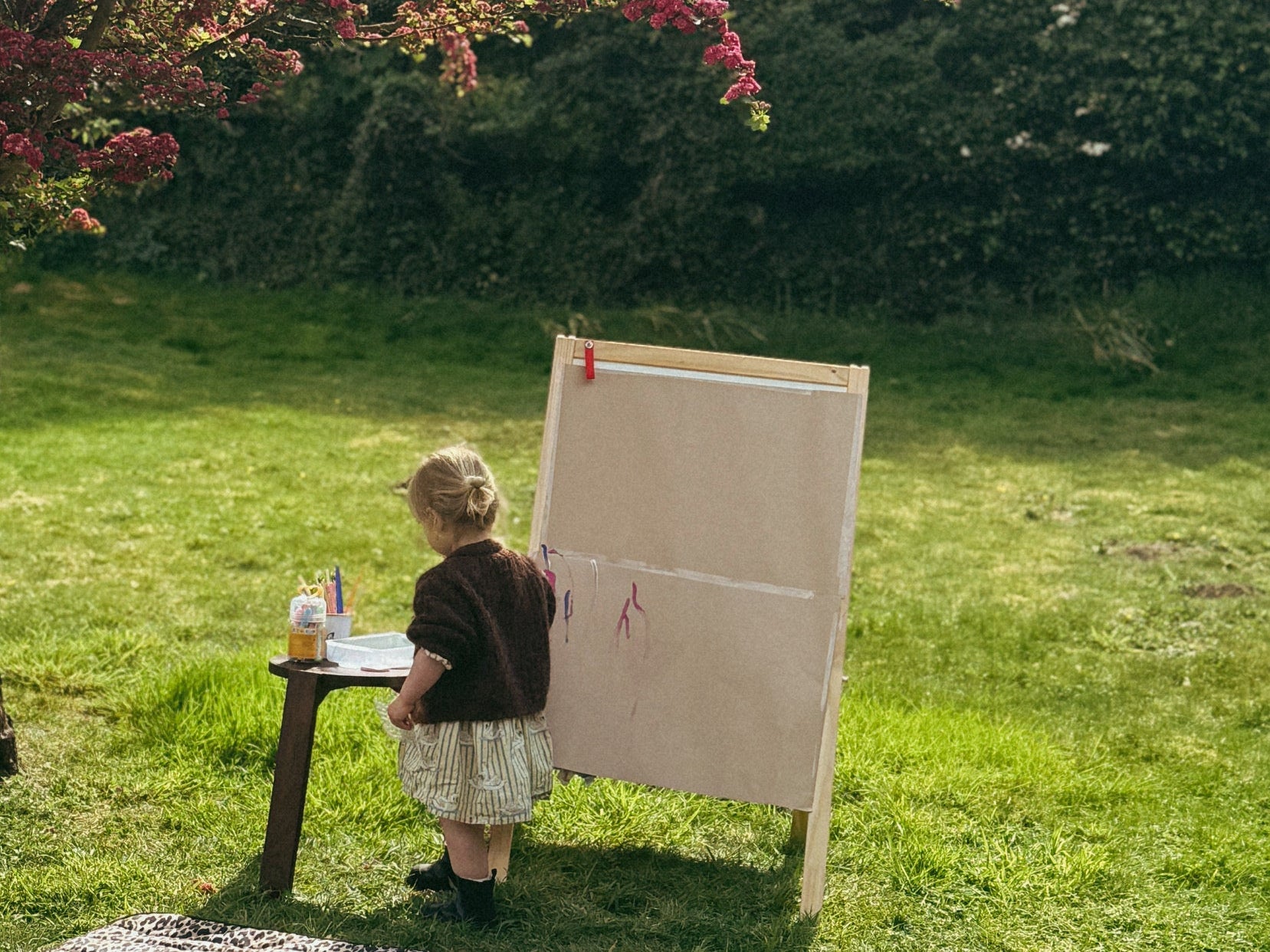 Thoren Eco Playmat in Leopard on a lawn with a baby lying on it while a toddler paints at an easel nearby