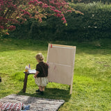 Thoren Eco Playmat in Leopard on a lawn with a baby lying on it while a toddler paints at an easel nearby