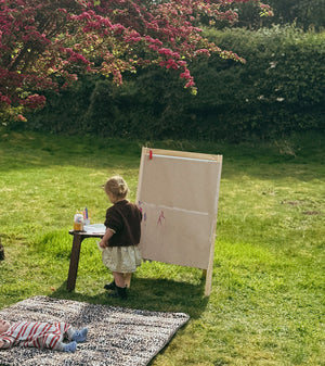 Thoren Eco Playmat in Leopard on a lawn with a baby lying on it while a toddler paints at an easel nearby