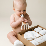 Sophie la girafe My Birth Outfit Gift Set with a baby chewing the giraffe toy beside an open box of white quilted items