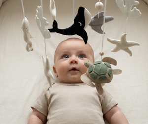 Ocean Whispers felt cot mobile hanging above a baby in a beige bodysuit lying in a cot, looking up at the felt sea animals