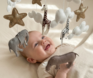 Safari Dreams Felt Cot Mobile hanging over a smiling baby lying on a cot, reaching up towards the felt rhino