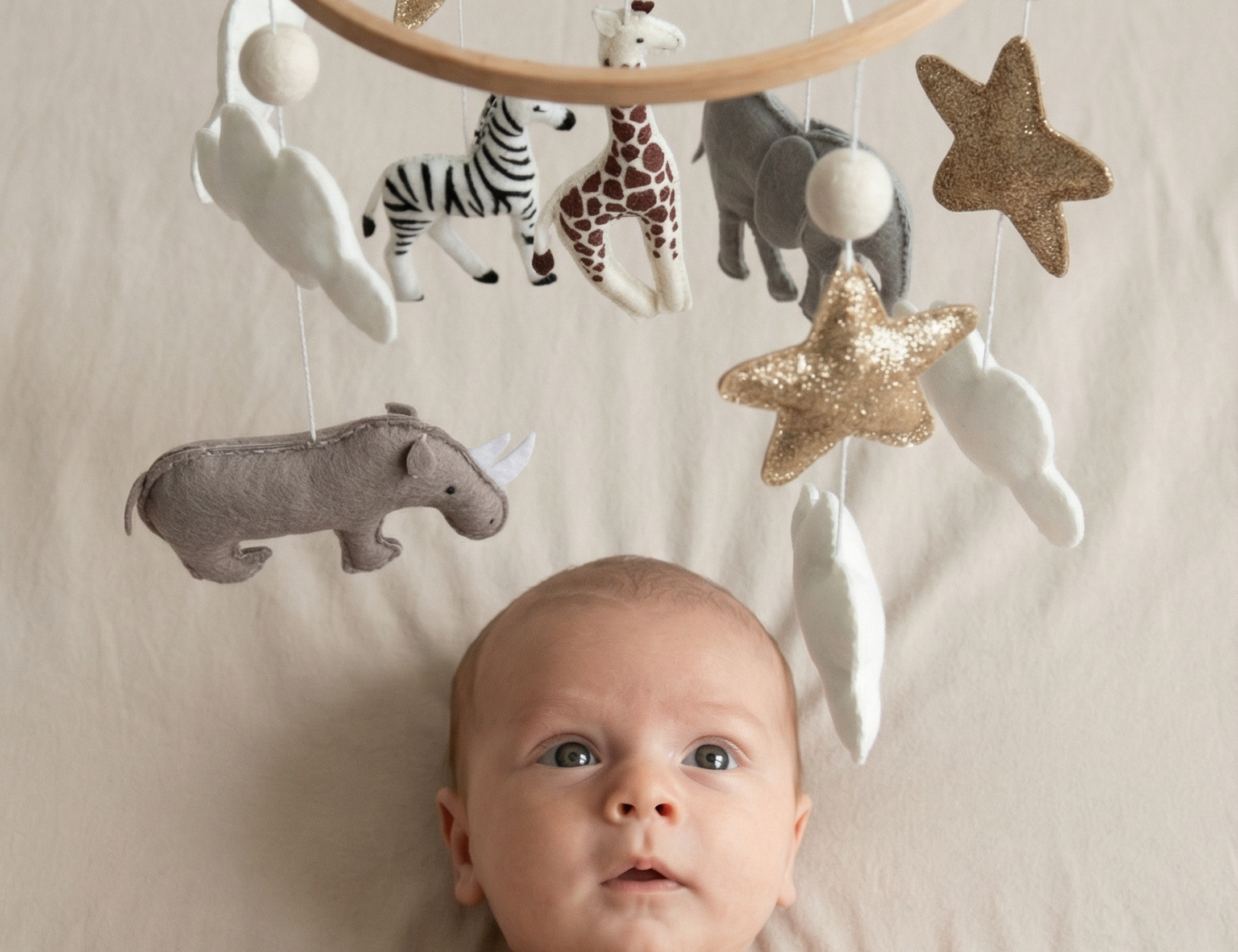 Safari Dreams Felt Cot Mobile above a wide-eyed baby lying on a bed, with the felt animals, stars and moon hanging