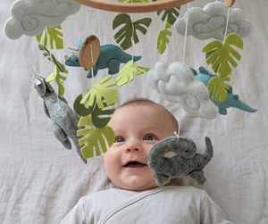 Little Dino Land felt cot mobile hanging over a smiling baby lying on their back in a cot
