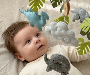 Little Dino Land felt cot mobile with a baby gazing up at the hanging blue and grey dinosaurs and clouds