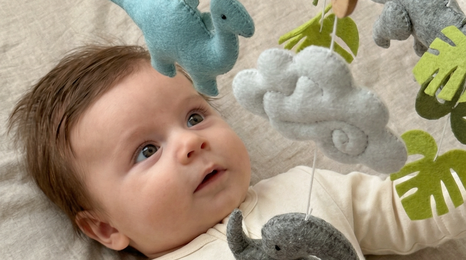 Little Dino Land felt cot mobile with a baby gazing up at the hanging blue and grey dinosaurs and clouds