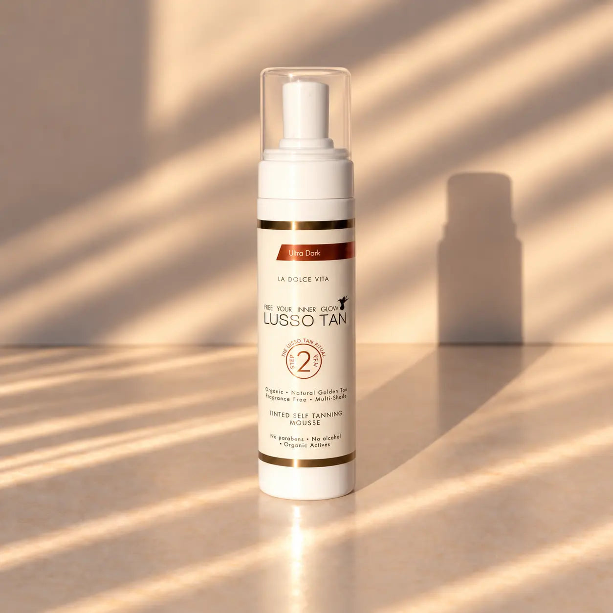 Lusso Tan tinted self tanning mousse, Ultra Dark bottle in warm sunlight with striped shadows on a beige surface