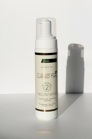 Lusso Tan tinted self tanning mousse, Medium shade bottle with green label stripe, standing in sunlight on a white surface