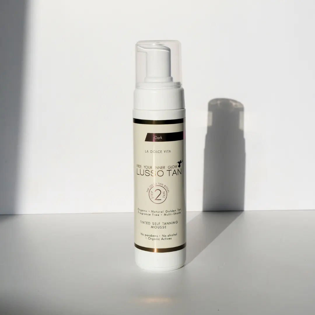 Lusso Tan tinted self tanning mousse, Dark shade bottle with pump top against a white wall with a soft shadow