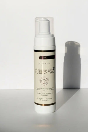 Lusso Tan tinted self tanning mousse, Dark shade bottle with pump top against a white wall with a soft shadow