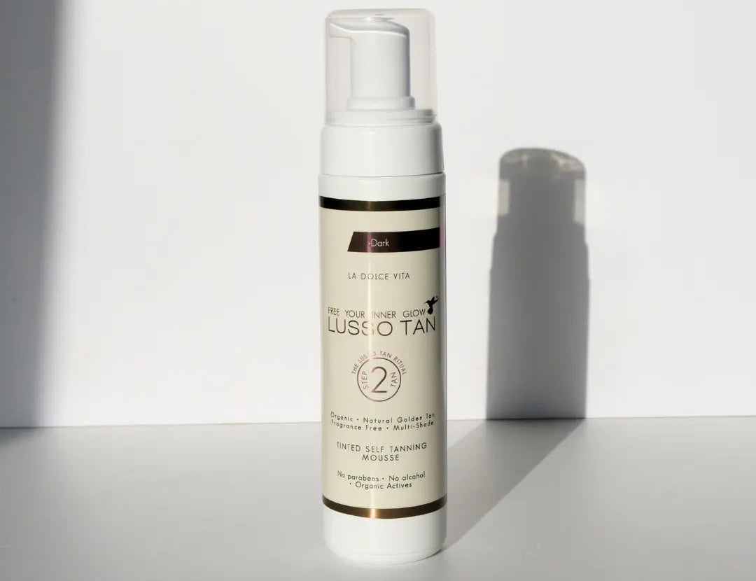 Lusso Tan tinted self tanning mousse, Dark shade bottle with pump top against a white wall with a soft shadow