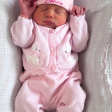 A sleeping newborn baby wearing a pink knit onesie, matching hat with bear ears, and white booties, lying on a textured white blanket. The hat is personalized with the name 'Bonnie' in pink embroidery.