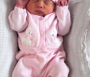 A sleeping newborn baby wearing a pink knit onesie, matching hat with bear ears, and white booties, lying on a textured white blanket. The hat is personalized with the name 'Bonnie' in pink embroidery.
