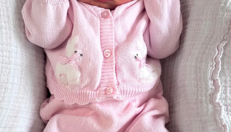 A sleeping newborn baby wearing a pink knit onesie, matching hat with bear ears, and white booties, lying on a textured white blanket. The hat is personalized with the name 'Bonnie' in pink embroidery.