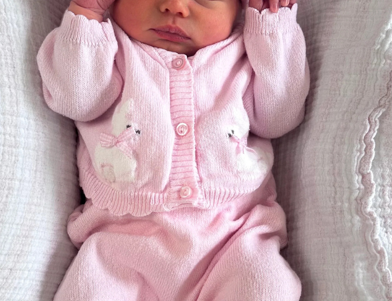 A sleeping newborn baby wearing a pink knit onesie, matching hat with bear ears, and white booties, lying on a textured white blanket. The hat is personalized with the name 'Bonnie' in pink embroidery.
