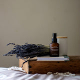 Calm Room Spray on a stack of books beside a bunch of dried lavender and a cream candle with a wooden lid