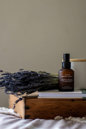 Calm Room Spray on a stack of books beside a bunch of dried lavender and a cream candle with a wooden lid