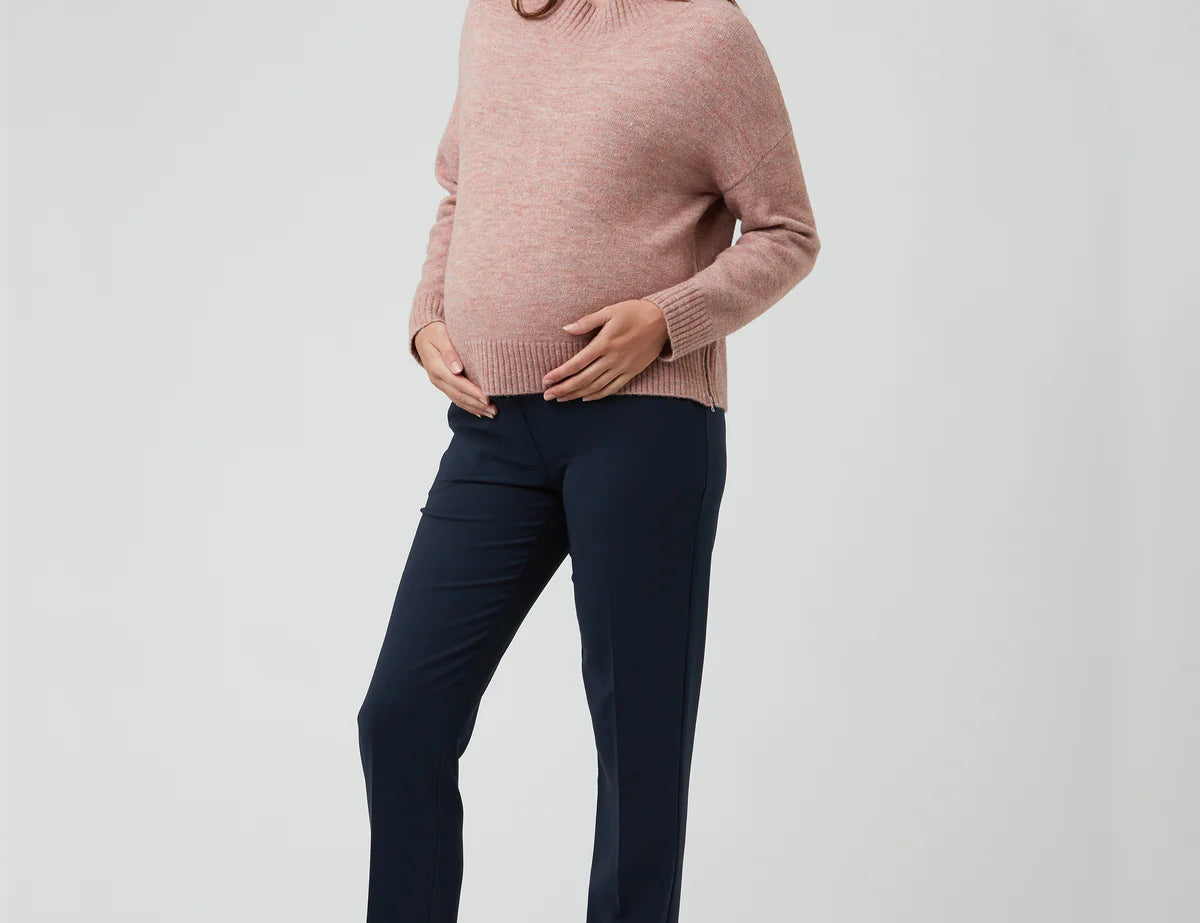Pregnant woman wearing a pink sweater and navy pants on a white background