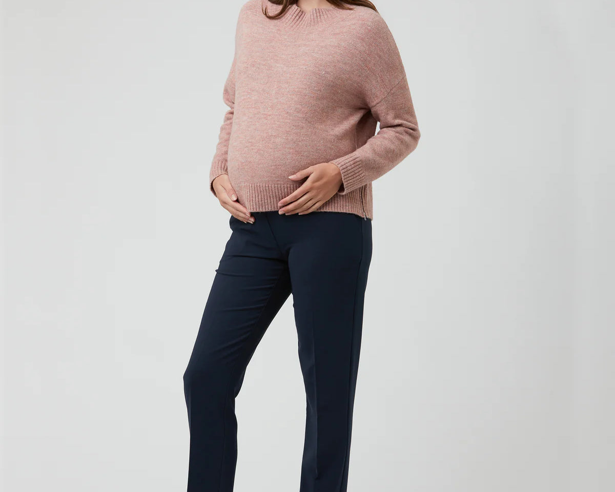 Pregnant woman wearing a pink sweater and navy pants on a white background